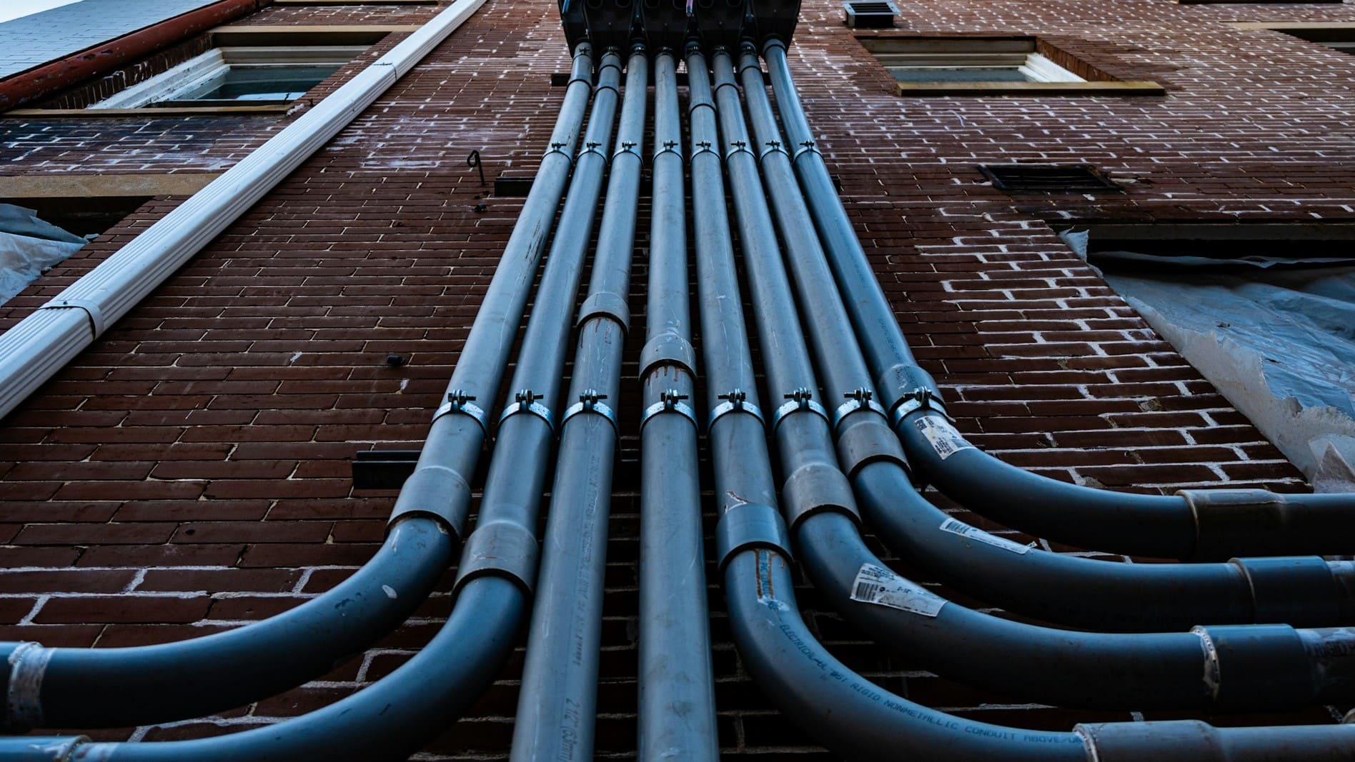 Leidingen verleggen tegen een bakstenen gevel met meerdere stevige grijze buizen die omhoog lopen